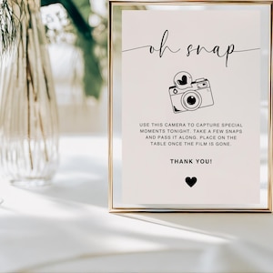 May include: A white sign with a gold frame and the text "oh snap" in black cursive font. A black and white illustration of a camera with a heart on the lens is above the text. The sign also includes the text "USE THIS CAMERA TO CAPTURE SPECIAL MOMENTS TONIGHT, TAKE A FEW SNAPS AND PASS IT ALONG. PLACE ON THE TABLE ONCE THE FILM IS GONE. THANK YOU!" in black text.