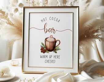 Könnte beinhalten: Ein gerahmtes Schild mit goldenem Rand auf einem weißen Tisch. Auf dem Schild steht "Hot Cocoa Bar" mit einer Aquarellillustration einer Tasse heißen Kakaos. Auf dem Schild steht auch "Warm Up Here Cheers!" Dekorative Pampasgras und weißer Stoff umgeben das Schild.