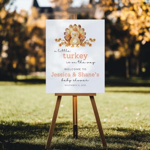 Puede incluir: Un caballete de madera exhibe un letrero blanco con una ilustraci&oacute;n de pavo y el texto "a little turkey is on the way". El letrero da la bienvenida a los invitados al baby shower de Jessica & Shane el 15 de noviembre de 2025. El fondo es una escena exterior borrosa.