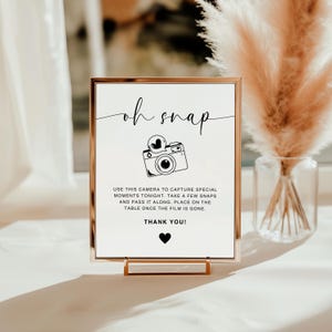 May include: A white sign with a gold frame and a black and white illustration of a camera. The sign reads "oh snap" and "USE THIS CAMERA TO CAPTURE SPECIAL MOMENTS TONIGHT, TAKE A FEW SNAPS AND PASS IT ALONG. PLACE ON THE TABLE ONCE THE FILM IS GONE. THANK YOU!"