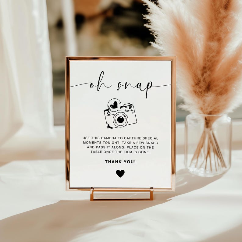 May include: A white sign with a gold frame and a black and white illustration of a camera. The sign reads "oh snap" and "USE THIS CAMERA TO CAPTURE SPECIAL MOMENTS TONIGHT, TAKE A FEW SNAPS AND PASS IT ALONG. PLACE ON THE TABLE ONCE THE FILM IS GONE. THANK YOU!"