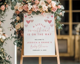 May include: A white sign with pink hearts and the text "A little sweetheart is on the way. Welcome to Jessica & Shane's Baby Shower. February 14, 20XX". The sign is on a wooden easel and decorated with pink flowers.