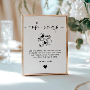 May include: A white sign with a gold frame that says "oh snap" in black cursive lettering. There is a black and white illustration of a camera with a heart above it. The sign also says "Use this camera to capture special moments tonight. Take a few snaps and pass it along. Place on the table once the film is gone. Thank you!"