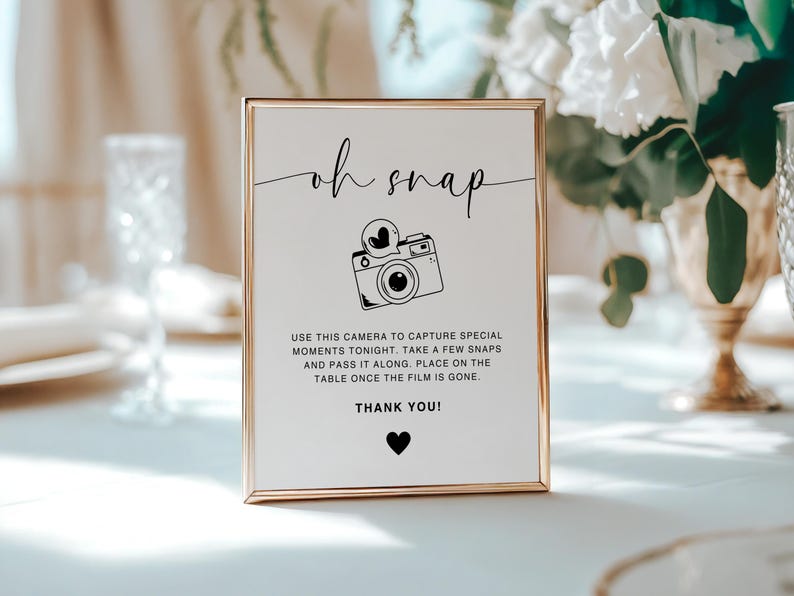 May include: A white sign with a gold frame that says "oh snap" in black cursive lettering. There is a black and white illustration of a camera with a heart above it. The sign also says "Use this camera to capture special moments tonight. Take a few snaps and pass it along. Place on the table once the film is gone. Thank you!"
