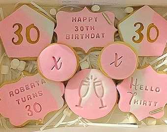 May include: A set of pink and white decorated sugar cookies with gold lettering. The cookies feature the text "Happy 30th Birthday", "Roberta Turns 30", "Hello Thirty", "30", and "x" and "t".