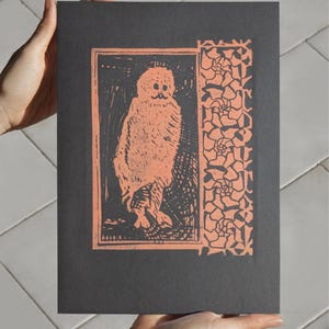 Peut inclure: Une impression noire et orange d'un hibou avec une bordure florale. Le hibou est représenté debout, les ailes repliées. L'impression est encadrée dans une bordure rectangulaire.