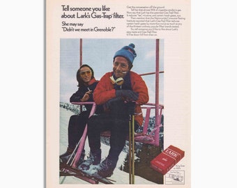May include: A vintage advert for Lark cigarettes featuring a man and woman on a ski lift. The man wears a red jacket and blue hat. The text reads, "Tell someone you like about Lark's Gas-Trap filter." The ad is from the 1970s.