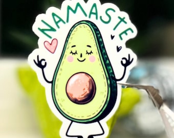 May include: A white sticker with a cartoon avocado meditating with the text "Namaste" above it. The avocado is green with a brown pit and has a smiling face with closed eyes. There are three pink hearts around the avocado.