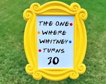 May include: A yellow picture frame with the text "THE ONE WHERE WHITNEY TURNS 30" on a white background. The frame is held up by a clear plastic stick. The background is green grass.