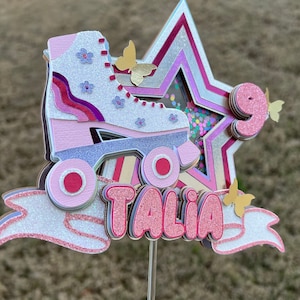 May include: A layered paper cake topper featuring a roller skate, star, and the name "TALIA" in pink glitter. The number "9" is also included. Butterflies and confetti add decorative elements. The color scheme is primarily pink, silver, and white.