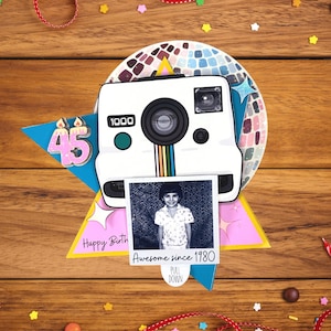 May include: A birthday card with a white camera design, a photo, and the number 45. The photo says "Awesome since 1980". The card is on a wooden surface with colorful confetti and ribbon.