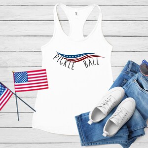 May include: White tank top with a red, white, and blue pickleball design and the text "Pickleball". The tank top is laying on a white wooden surface with two small American flags, a pair of blue jeans, and white sneakers.