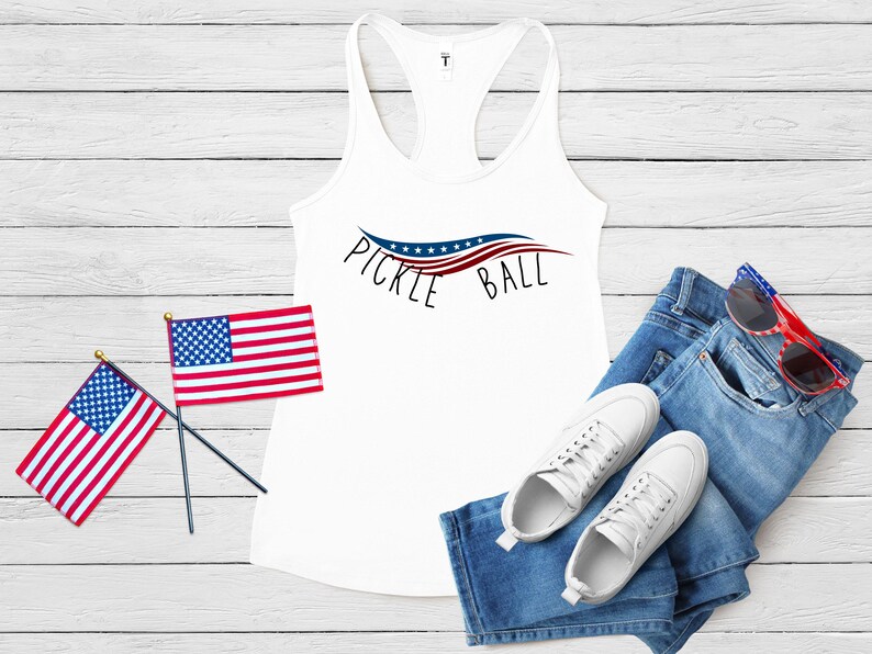May include: White tank top with a red, white, and blue pickleball design and the text "Pickleball". The tank top is laying on a white wooden surface with two small American flags, a pair of blue jeans, and white sneakers.