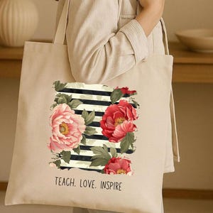 Puede incluir: Bolso tote beige con un dise&ntilde;o floral de peon&iacute;as rosas y rojas, hojas verdes y rayas blancas y negras. El texto "TEACH. LOVE. INSPIRE" est&aacute; impreso en la parte inferior. Un accesorio elegante y pr&aacute;ctico.