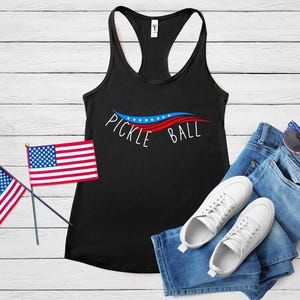 May include: Black tank top with a red, white, and blue wave design and the text "Pickle Ball". The tank top is paired with blue jeans, white sneakers, and red, white, and blue sunglasses.