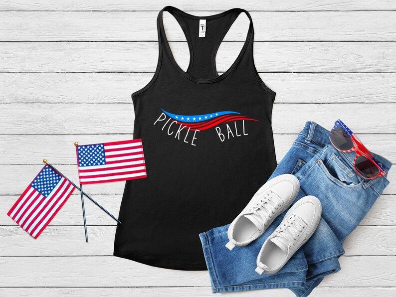 May include: Black tank top with a red, white, and blue wave design and the text "Pickle Ball". The tank top is paired with blue jeans, white sneakers, and red, white, and blue sunglasses.