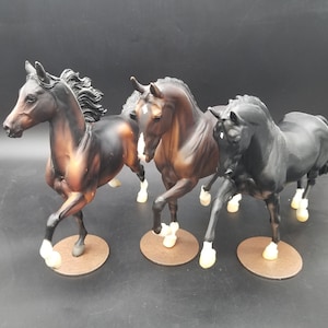 May include: Three horse figurines, one brown and white, one black and white, and one brown and black. The horses are standing on a black surface.