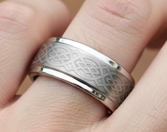 May include: A silver-colored ring with a Celtic knot design. The ring is wide with a polished finish and a detailed pattern etched into the band. The ring is on a finger.