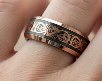 May include: A rose gold ring with a black band featuring an intricate Celtic knot design. The ring is shown on a finger, showcasing the detailed pattern and metallic finish. The ring's design is a blend of modern and traditional aesthetics.