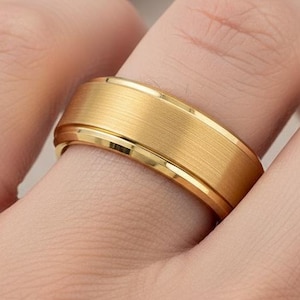 May include: A gold-colored ring with a brushed, textured center band and polished edges. The ring is on a finger, showcasing its design and metallic sheen. The ring's width is approximately 0.3 inches.