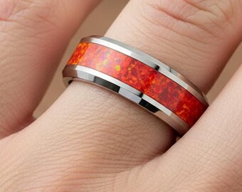 May include: A silver ring with a vibrant red-orange center stone. The ring is on a finger, showcasing the polished metal band and the fiery color of the stone. The ring's design is modern and eye-catching, suitable for everyday wear.