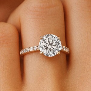 May include: A gold ring with a large, round, clear gemstone as the centerpiece. The band is also adorned with smaller, clear gemstones. The ring is on a person's finger, with the hand and part of the face visible.