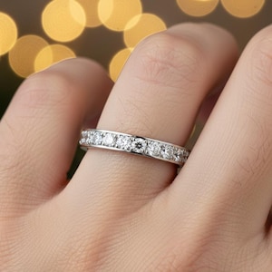 May include: A silver band ring with multiple small, clear gemstones set around the entire band. The ring is on a person's finger, with a blurred background of golden bokeh lights.