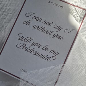 May include: A white card with burgundy trim and elegant script, asking "Will you be my Bridesmaid?" The card rests on a white, textured fabric, creating a soft, romantic aesthetic. The text reads "I can not say I do, without you."