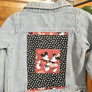 May include: Light blue denim jacket with a Mickey and Minnie Mouse patch on the back. The patch features a black and white polka dot pattern with a red border and a red square with Mickey and Minnie Mouse.