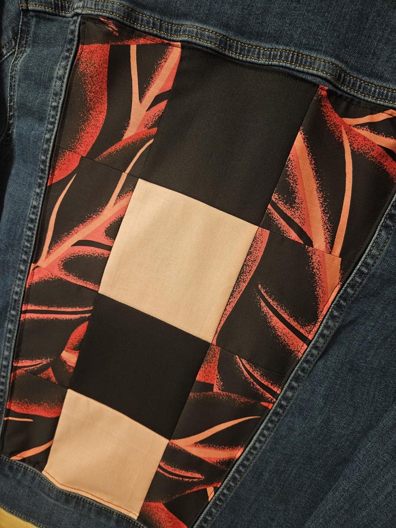 Puede incluir: Una chaqueta de mezclilla azul oscuro con un panel trasero rectangular hecho de un dise&ntilde;o de patchwork. El panel consta de cuadrados negros s&oacute;lidos, cuadrados beige s&oacute;lidos y tela negra con un patr&oacute;n de hojas rojas y naranjas.