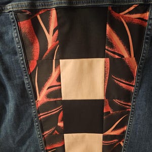 Puede incluir: Una chaqueta de mezclilla azul con un panel central grande en la espalda hecho con un dise&ntilde;o de patchwork. El panel combina cuadrados negros s&oacute;lidos, cuadrados beige s&oacute;lidos y cuadrados de tela con un estampado de hojas tropicales negro y naranja.