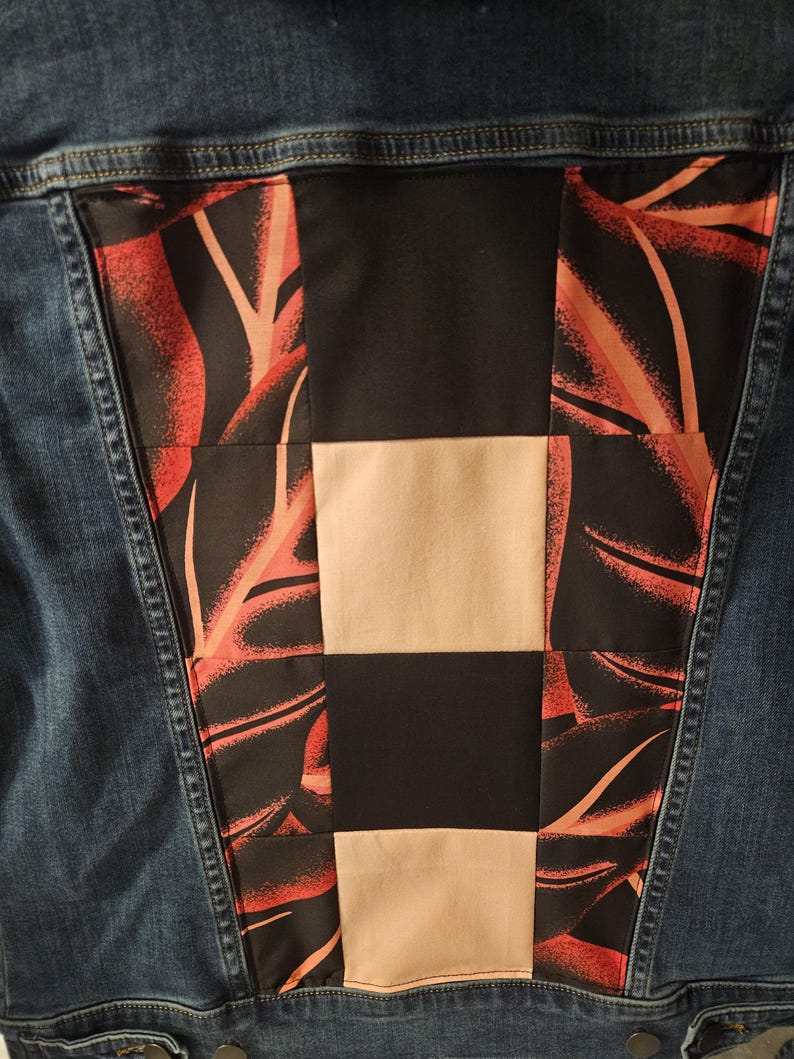 Puede incluir: Una chaqueta de mezclilla azul con un panel central grande en la espalda hecho con un dise&ntilde;o de patchwork. El panel combina cuadrados negros s&oacute;lidos, cuadrados beige s&oacute;lidos y cuadrados de tela con un estampado de hojas tropicales negro y naranja.
