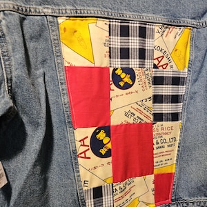 May include: Light blue denim jacket with a patchwork design on the back. The patchwork includes red squares, yellow and white patterned fabric with text, and black and white plaid fabric. The jacket has a classic collar and button closure.