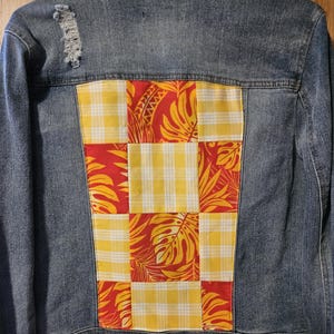 May include: A blue denim jacket with a red and yellow patchwork design on the back. The patchwork is made up of squares of red fabric with a yellow and white plaid pattern and yellow fabric with a red and orange floral pattern.