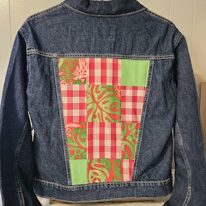 May include: A dark blue denim jacket featuring a large, rectangular patchwork panel on the back. The panel consists of alternating squares of red and white plaid fabric, bright green fabric with a red leaf pattern, and solid light green fabric.