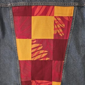 May include: Close-up of a denim jacket with a patchwork design on the back. The patchwork features a combination of red, yellow, and orange squares and leaf patterns, creating a unique and colorful accent.
