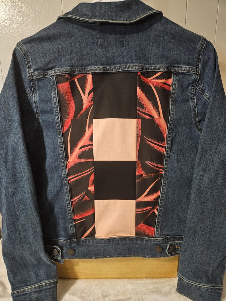 Puede incluir: Una chaqueta de mezclilla azul con un panel trasero de dise&ntilde;o patchwork. El panel consta de cuadrados alternos de tela negra s&oacute;lida, tela rosa claro s&oacute;lida y tela negra con un patr&oacute;n de hojas rojas, cosidos en la parte posterior de mezclilla.