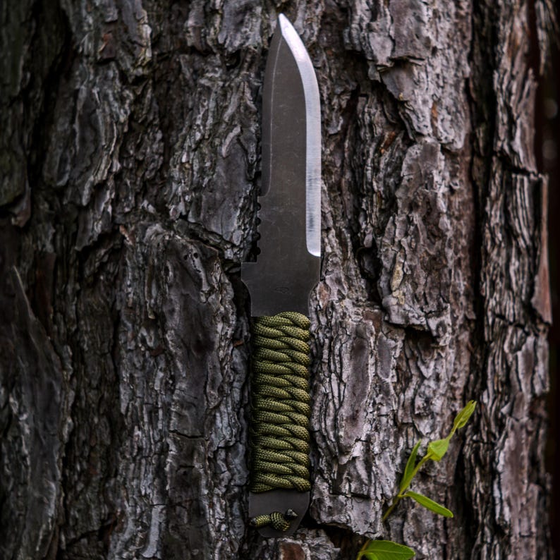 May include: A fixed-blade knife with a dark gray metal blade and a handle wrapped in olive green paracord. The blade features a straight edge and a pointed tip, while the handle includes a small lanyard loop at the base for secure handling.