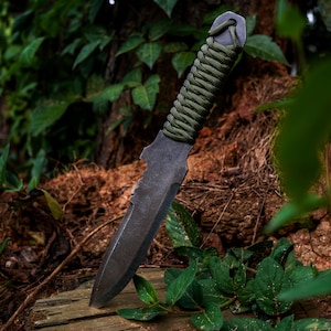 May include: A dark gray metal knife with a pointed tip and a serrated edge near the handle. The handle is wrapped in olive green paracord, and the letters CM are stamped into the metal blade near the hilt, featuring a lanyard hole at the end.