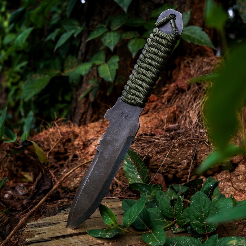 May include: A dark gray metal knife with a pointed tip and a serrated edge near the handle. The handle is wrapped in olive green paracord, and the letters CM are stamped into the metal blade near the hilt, featuring a lanyard hole at the end.