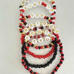 May include: A collection of seven elastic beaded bracelets featuring red, black, white, and gold beads. Several bracelets include white letter beads with gold text spelling out WRECK EM, GUNS UP, LBK, RAIDER, POWER, and TTU, arranged in a stacked formation.