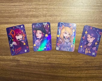 May include: Four holographic cards featuring different characters from the video game League of Legends. The cards are purple with pink and yellow accents. The characters are Evelynn, Ahri, Kai'Sa, and Akali. Each card has the character's name printed on it.