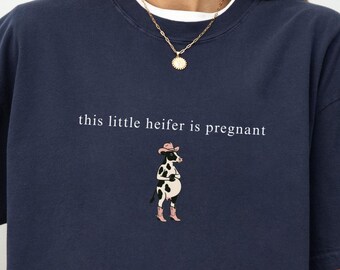 Może przedstawiać: Granatowa koszulka z napisem "this little heifer is pregnant" i ilustracją krowy w stroju kowbojskim. Koszulka jest w kolorze granatowym.