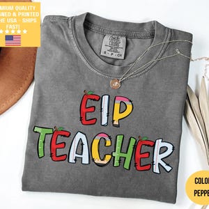以下が含まれることがあります： カラフルで遊び心のある文字で「EIP TEACHER」と書かれたグレーのComfort Colors Tシャツ。クルーネックと半袖です。ペンダント付きのゴールドネックレスがシャツの上にかけられています。シャツの色はペッパーです。