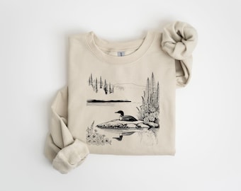 May include: A beige sweatshirt with a black and white illustration of a loon on a lake with trees and mountains in the background.