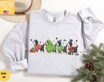 Puede incluir: Sudadera gris ceniza con un diseño festivo de cuatro perros de montaña berneses vestidos con atuendos navideños. Un perro lleva un disfraz de árbol de Navidad, mientras que otros lucen suéteres y un atuendo de cascanueces. Los regalos y adornos completan el tema.
