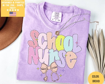 May include: A light purple t-shirt with the words "School Nurse" in a colorful, checkered font. The design includes a bow, stethoscope, and stars. The shirt is labeled "Comfort Colors" and the color is "Orchid".