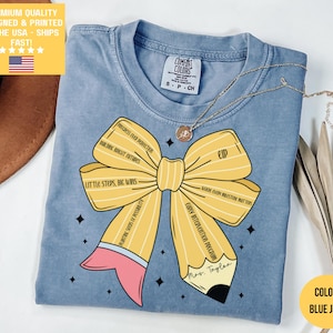 May include: Blue jean coloured t-shirt featuring a yellow pencil design. The pencil is shaped like a bow with text that reads "Progress over perfection", "Build bright futures", "Little steps, big wins", and "Planting seeds of possibility".
