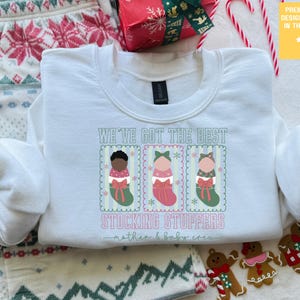 May include: White sweatshirt with the text "WE'VE GOT THE BEST STOCKING STUFFERS mother & baby crew." The graphic features three baby stocking designs. The sweatshirt is surrounded by Christmas decorations.