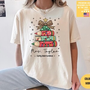 May include: Ivory-colored t-shirt featuring a Christmas tree design made of pencils and books, adorned with a leopard print bow and star. The text "Mrs. Taylor Early Intervention" is printed below the tree. The shirt has short sleeves.